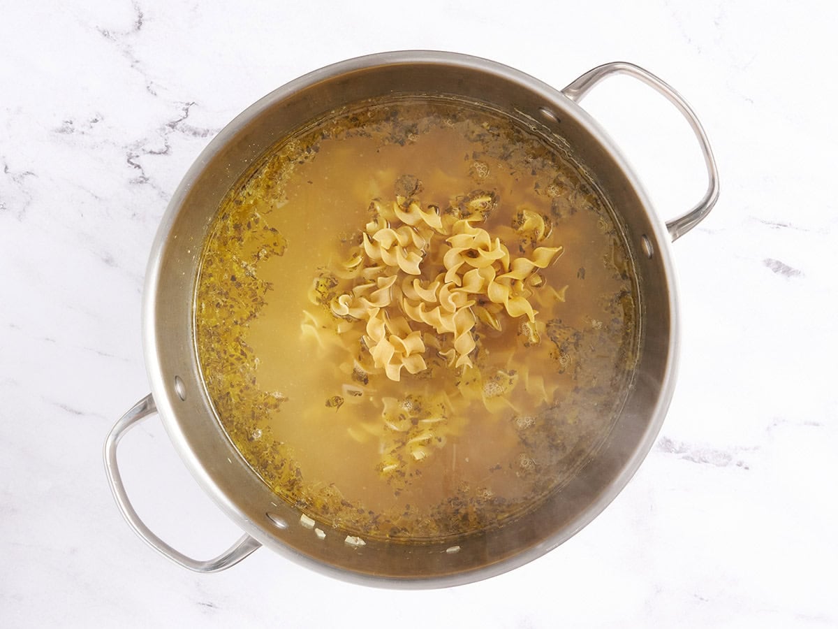 Uncooked egg noodles added to chicken noodle soup in a soup pot.