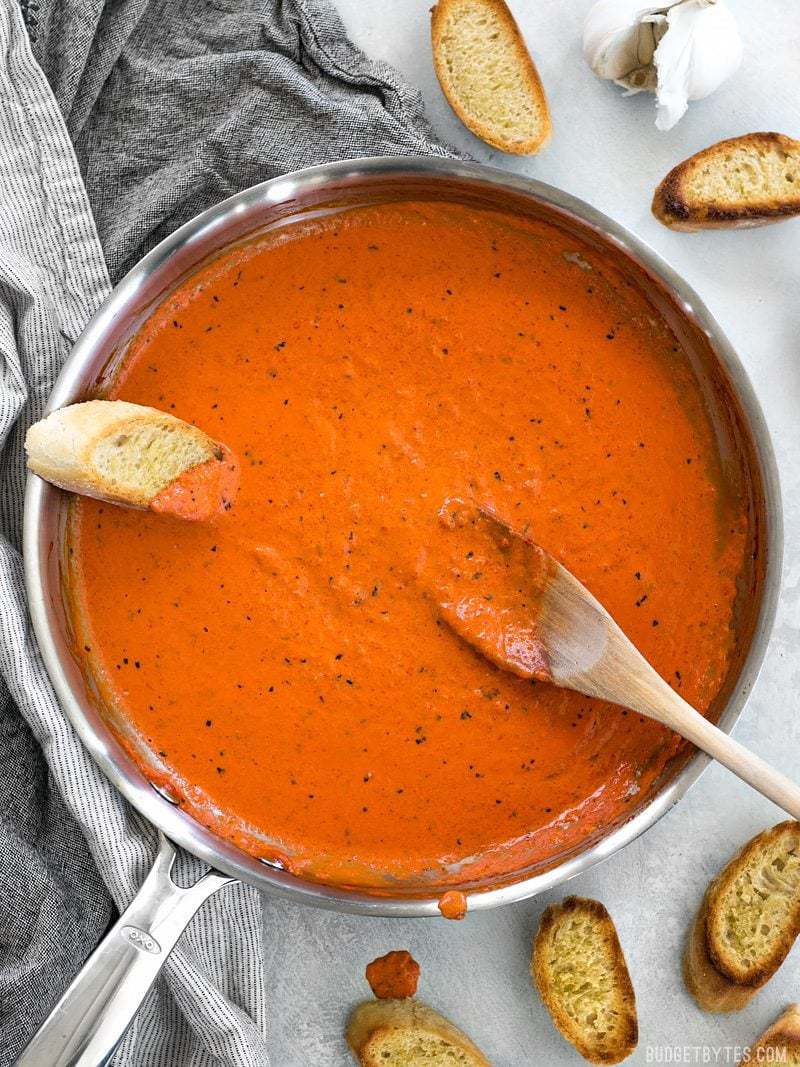 Creamy Roasted Red Pepper Sauce is a great alternative to tomato based sauces for pasta, pizzas, or just for dipping your favorite crusty bread. BudgetBytes.com Creamy Roasted Red Pepper Sauce in a skillet with slices of bread and a wooden spoon