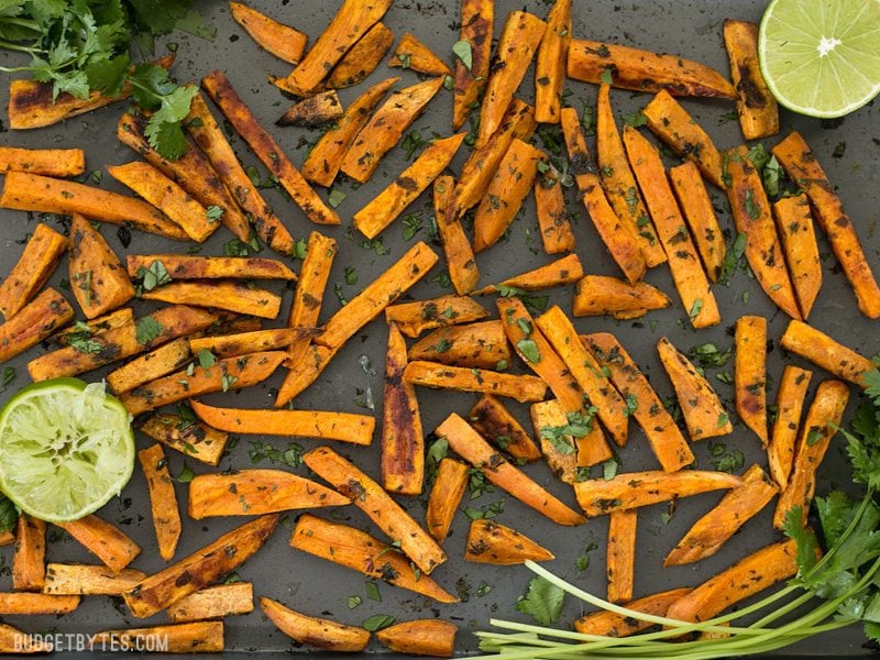 Bright lime juice and earthy cumin pair perfectly with the subtle creamy sweetness of sweet potatoes in these Cumin Lime Roasted Sweet Potatoes. BudgetBytes.com Finished Cumin Lime Roasted Sweet Potatoes on the baking sheet with cilantro and limes