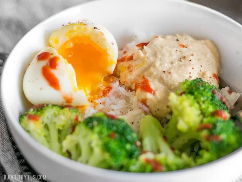 Hummus Breakfast Bowls are a medley of colors, flavors, and textures, and a great way to work vegetables into the most important meal of the day. BudgetBytes.com Close up side view of a Hummus Breakfast Bowl with the soft boiled egg cracked open and spilling yolk