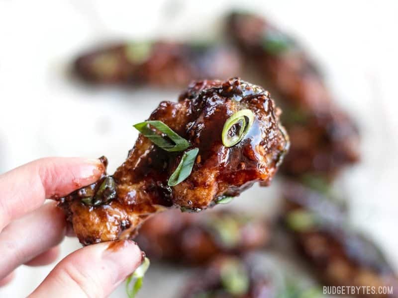 These crispy Raspberry Balsamic Baked Chicken Wings are baked, not fried, and slathered in a sweet, tangy, and rich homemade sauce. BudgetBytes.com Close up of one Raspberry Balsamic Baked Chicken Wing being held in a hand, sauce getting on the fingers