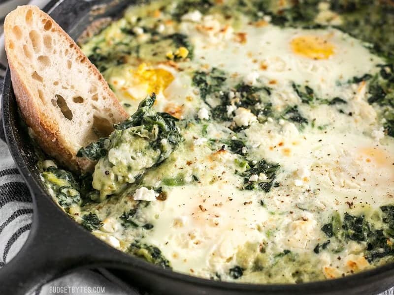 Make these fast and easy Creamed Spinach Baked Eggs using items you probably have in your pantry. A little feta on top takes it to the next level! BudgetBytes.com Close up shot of a piece of bread being dipped into Creamed Spinach Baked Eggs