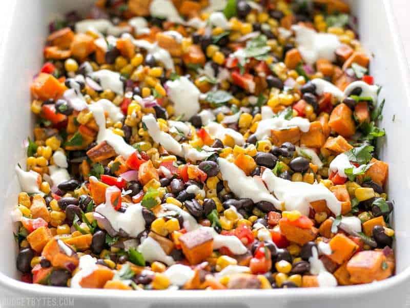 This Roasted Sweet Potato Rainbow Salad combines a medley of vibrant colors and flavors, brought together by a bright and creamy dressing. BudgetBytes.com Roasted Sweet Potato Rainbow Salad in a large serving dish, from the front, with lime crema drizzled on top