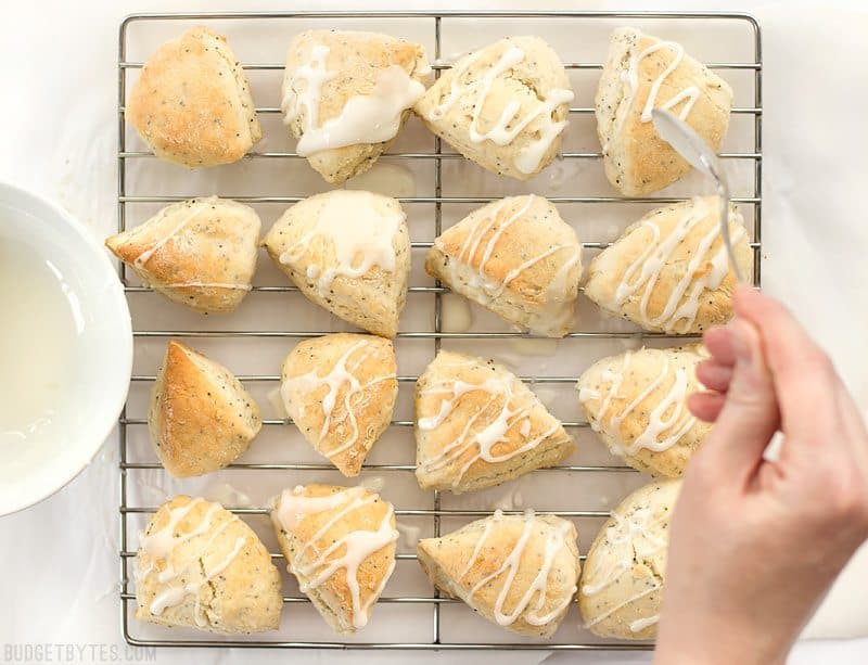 These cute little lemon poppy seed two-bite scones are soft and sweet with a deliciously tart lemon glaze. You won't be able to have just one! BudgetBytes.com Lemon glaze being drizzled over lemon poppy seed two-bite scones on a wire cooling rack
