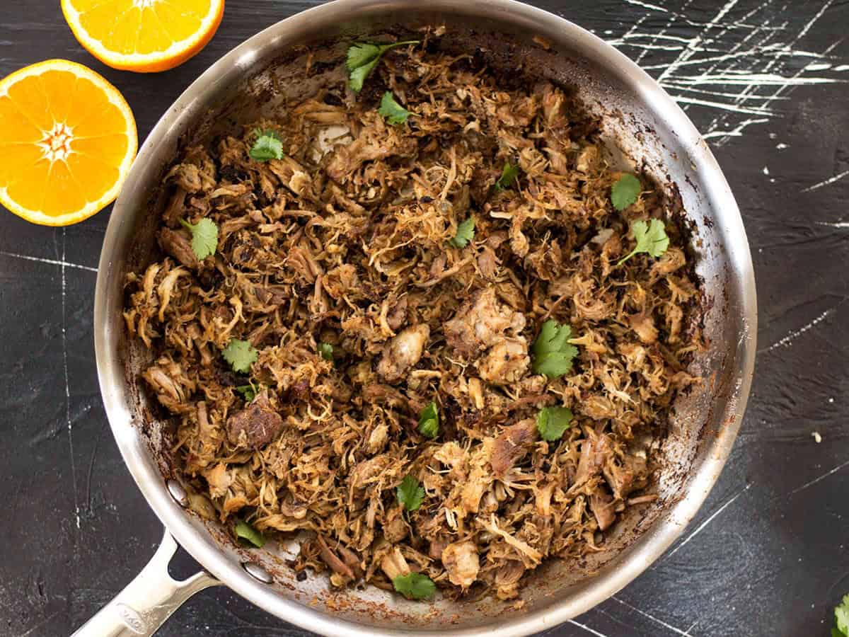 Warm spices, fresh orange essence, and a low slow cook time makes this Slow Cooker Carnitas tender, juicy, and full of flavor. Perfect for tacos, burritos and more! Overhead view of a skillet full of slow cooker carnitas with an orange on the side.