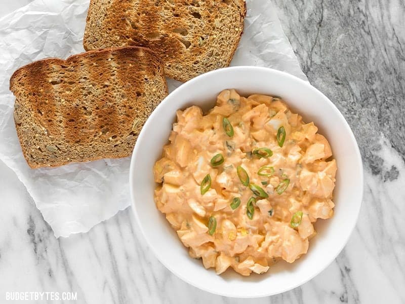 Sriracha Egg Salad is a simple yet satisfying dish that boasts a creamy, tangy, and spicy sauce. BudgetBytes.com A bowl of Sriracha Egg Salad next to two slices of toast