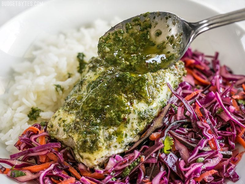 These light and fresh Baked Chimichurri Fish Bowls are simple to prepare and make great cold lunches for the rest of the week. BudgetBytes.com Chimichurri being drizzled over a piece of baked fish sitting on top of red cabbage slaw