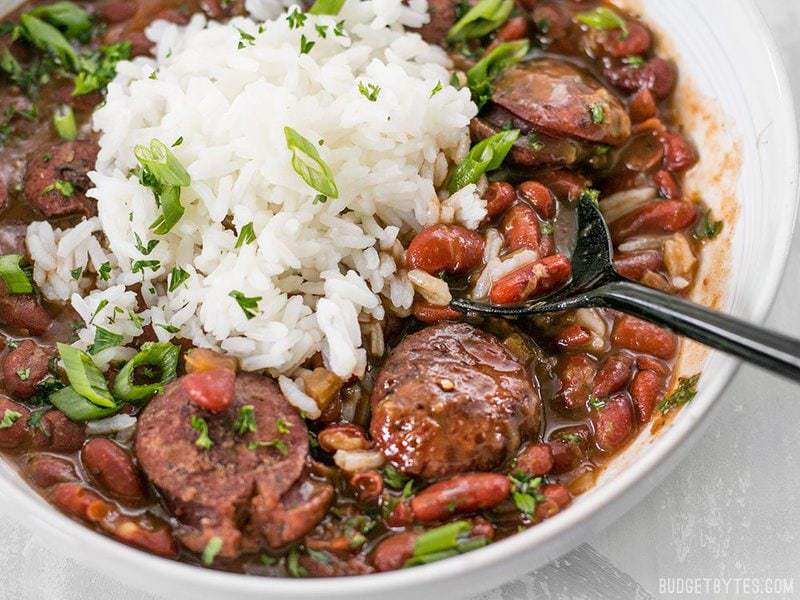 Pressure Cooker Red Beans are a fast, inexpensive, filling, and flavorful staple meal. It's a Louisiana tradition, but FASTER! BudgetBytes.com Close up side view of a bowl of Pressure Cooker Red Beans with a black spoon lifting a scoop