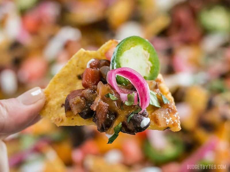 For the perfect Spicy Baked Black Bean Nachos, layer your chips and toppings for the perfect chip-to-topping ratio. BudgetBytes.com Close up of a Spicy Baked Black Bean Nacho with pickled onions and a slice of jalapeno