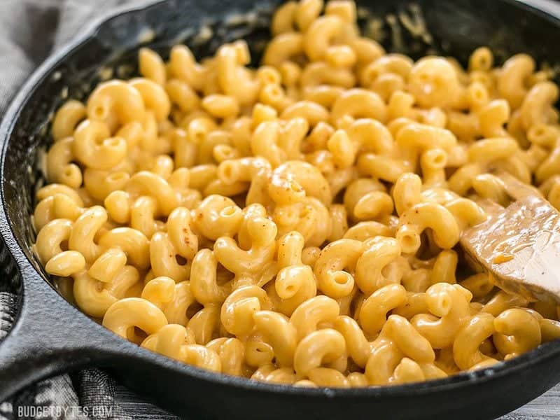 This incredible roux-less mac and cheese is rich, creamy, and only requires seven ingredients. Perfect for last minute weeknight dinners! BudgetBytes.com Close up side view of a cast iron skillet full of mac and cheese, with a wooden spoon