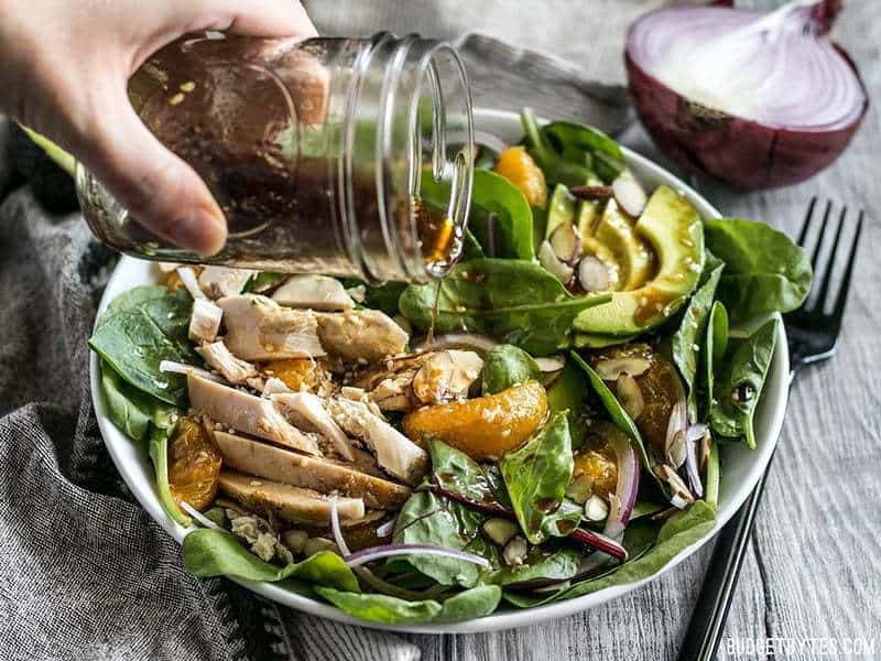 This meal-worthy Chicken and Mandarin Salad boasts sweet mandarin oranges, creamy avocado, crunchy almonds, and homemade sesame dressing. BudgetBytes.com Sesame dressing being poured from a mason jar onto Chicken and Mandarin Salad