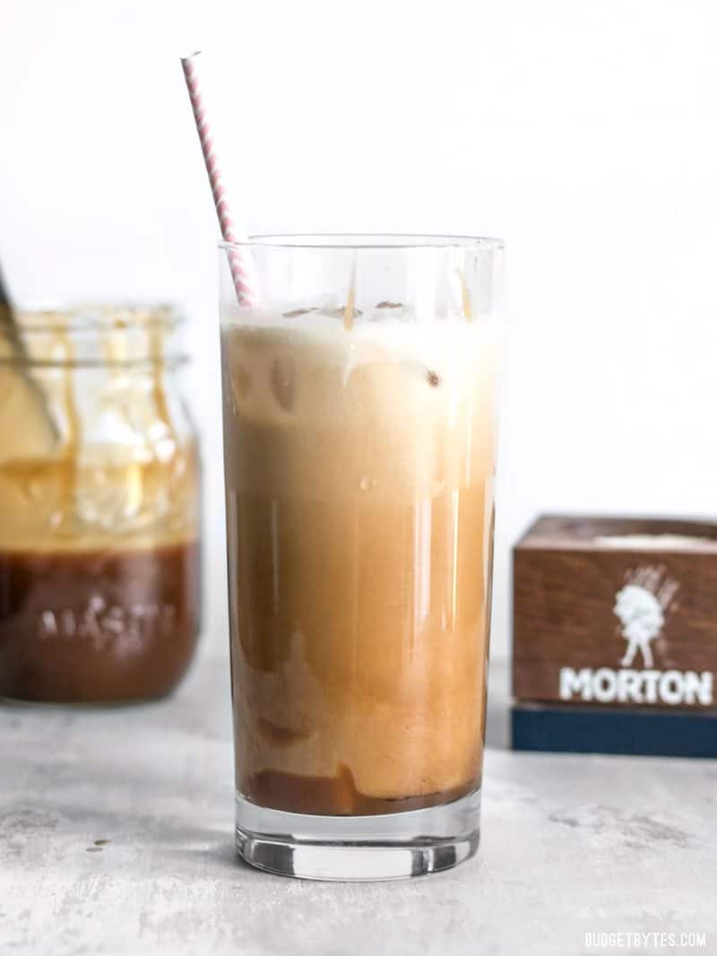 A little fine sea salt helps this Homemade Salted Caramel Iced Coffee stay smooth and taste extra sweet. Make this coffee house favorite at home for less! #ad BudgetBytes.com Homemade Salted Caramel Iced Coffee viewed from the side with a jar full of caramel and a salt conter in the background