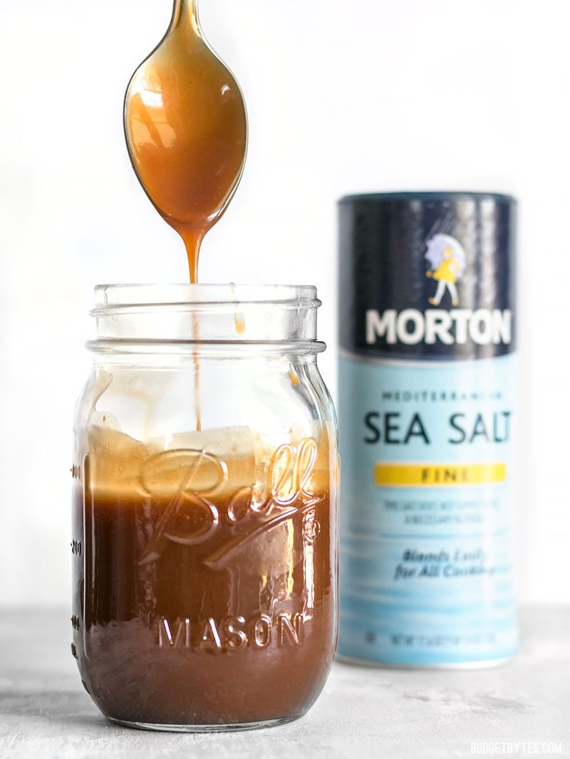 A little fine sea salt helps this Homemade Salted Caramel Iced Coffee stay smooth and taste extra sweet. Make this coffee house favorite at home for less! #ad BudgetBytes.com Homemade caramel dripping off a spoon into a mason jar with salt container in the background