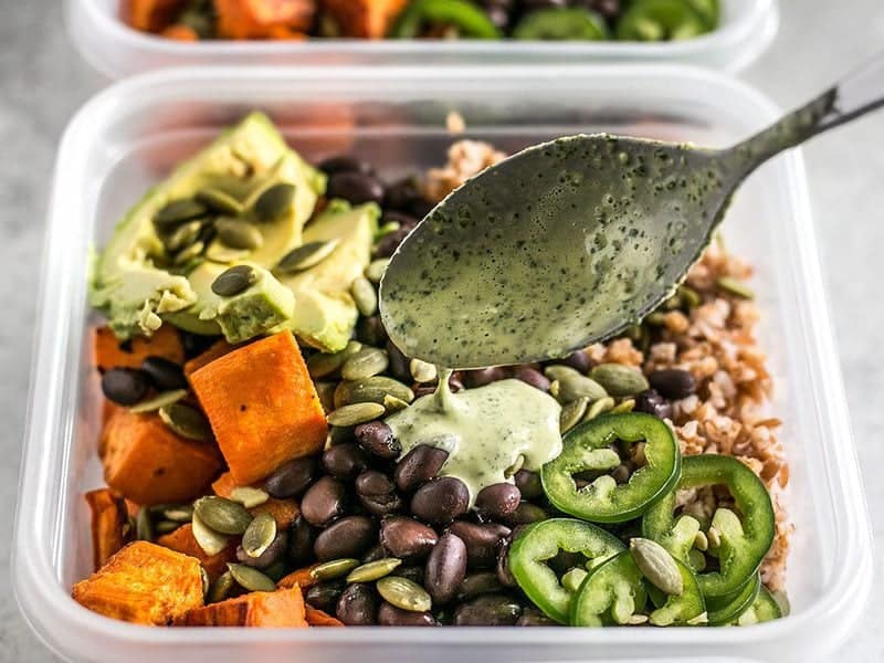 These Sweet Potato Grain Bowls with Green Tahini Sauce are prefect for meal prep and bursting with color, texture, and flavor! BudgetBytes.com Green Tahini Sauce being drizzled over a meal prep container full of sweet potato grain bowls