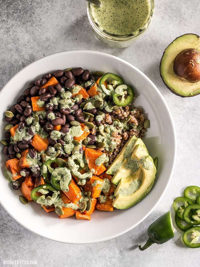These Sweet Potato Grain Bowls with Green Tahini Sauce are prefect for meal prep and bursting with color, texture, and flavor! BudgetBytes.com A prepared Sweet Potato Grain Bowl with a jar of green tahini dressing on the side