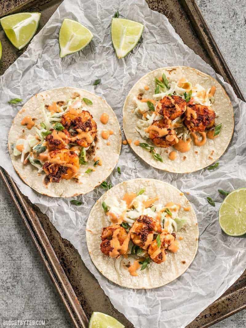 Smoky and spicy shrimp, sweet and tangy slaw, and a zesty garlic lime sauce make these Blackened Shrimp Tacos seriously delicious! Three open Blackened Shrimp Tacos on a metal baking sheet with lime wedges nearby