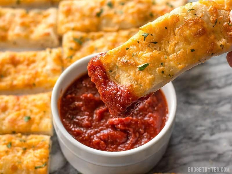 This Homemade Cheesy Garlic Bread is made from scratch with homemade dough, fresh garlic, real butter, and your favorite cheese. Budgetbytes.com One piece of Homemade Cheesy Garlic Bread being dipped into pizza sauce