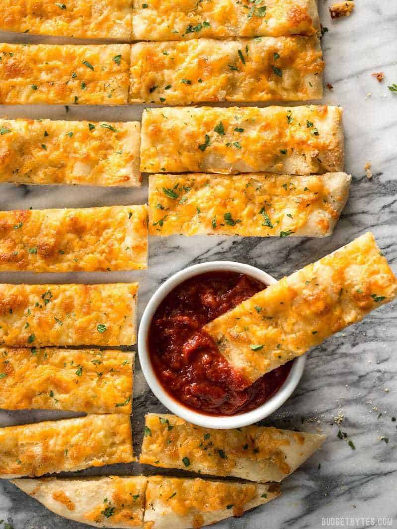 This Homemade Cheesy Garlic Bread is made from scratch with homemade dough, fresh garlic, real butter, and your favorite cheese. Budgetbytes.com Overhead view of sliced Homemade Cheesy Garlic Bread with a dish of pizza sauce