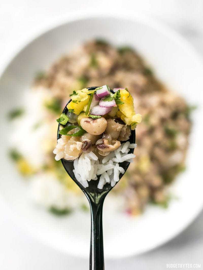 These rich and spicy Coconut Jerk Peas are super simple to make and pair brilliantly with a sweet and vibrant pineapple salsa. BudgetBytes.com Close up of a spoonful of Coconut Jerk Peas with rice and pineapple salsa