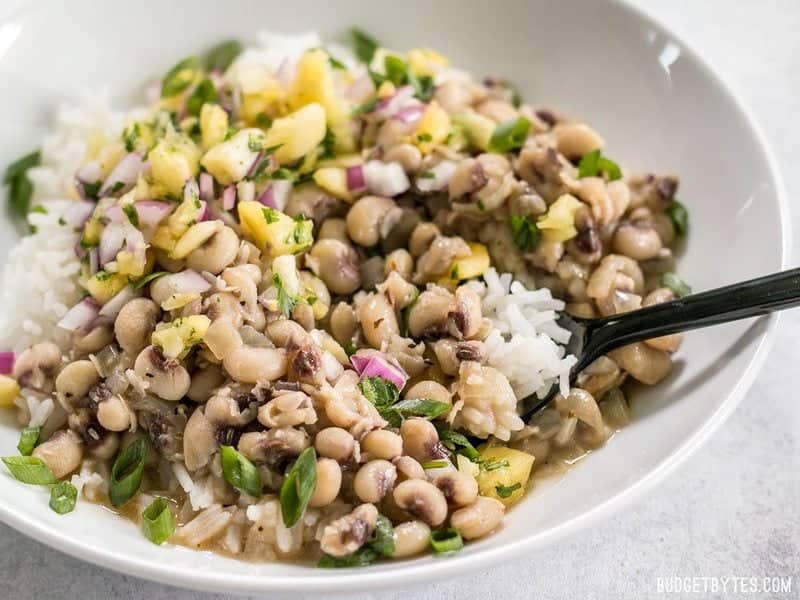 These rich and spicy Coconut Jerk Peas are super simple to make and pair brilliantly with a sweet and vibrant pineapple salsa. BudgetBytes.com Close up side view of a bowl of Coconut Jerk Peas with rice and pineapple salsa