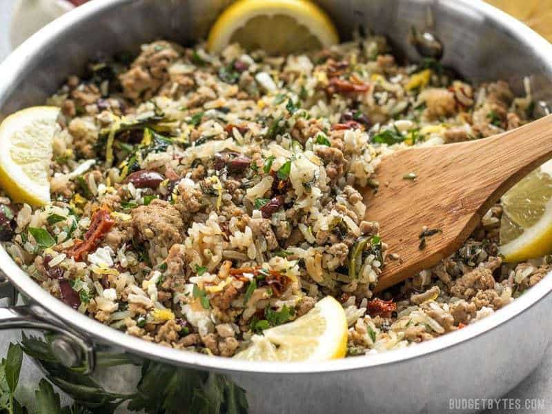 Greek Turkey and Rice Skillet is an easy and flavorful ground turkey recipe that cooks in one skillet for maximum flavor and minimum effort. Budgetbytes.com A wooden spatula stirring the Greek Turkey and Rice Skillet, with lemon wedges on the sides.