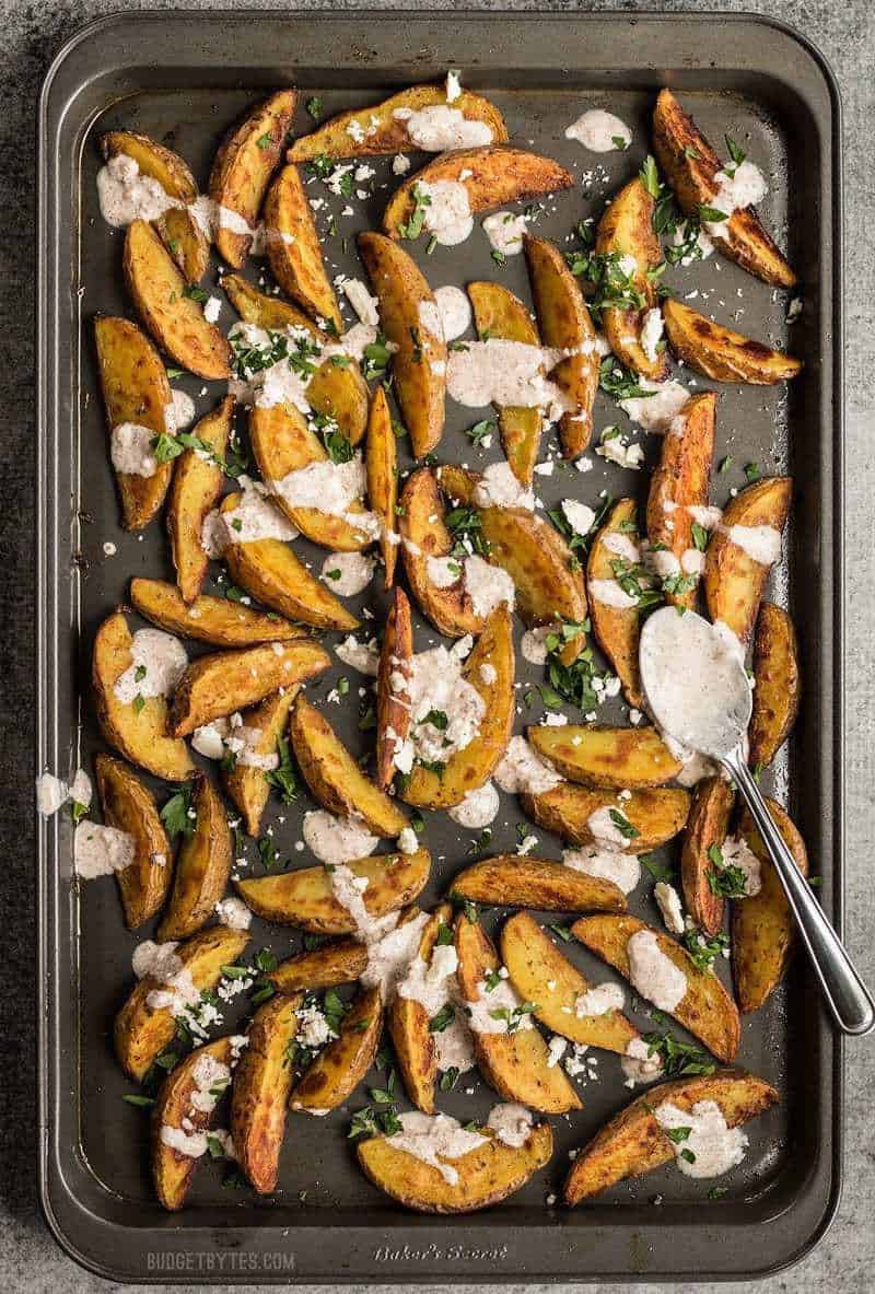 These simple Roasted Potato Wedges are the perfect vehicle for this creamy, garlicky shawarma sauce and crumbled feta. BudgetBytes.com Roasted Potato Wedges with shawarma sauce on the baking sheet with a spoon full of shawarma sauce