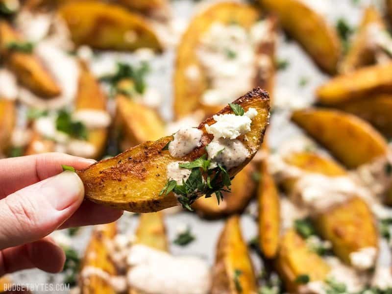 These simple Roasted Potato Wedges are the perfect vehicle for this creamy, garlicky shawarma sauce and crumbled feta. BudgetBytes.com Close up of one Roasted Potato Wedge held in a hand, with the sheet pan in the background