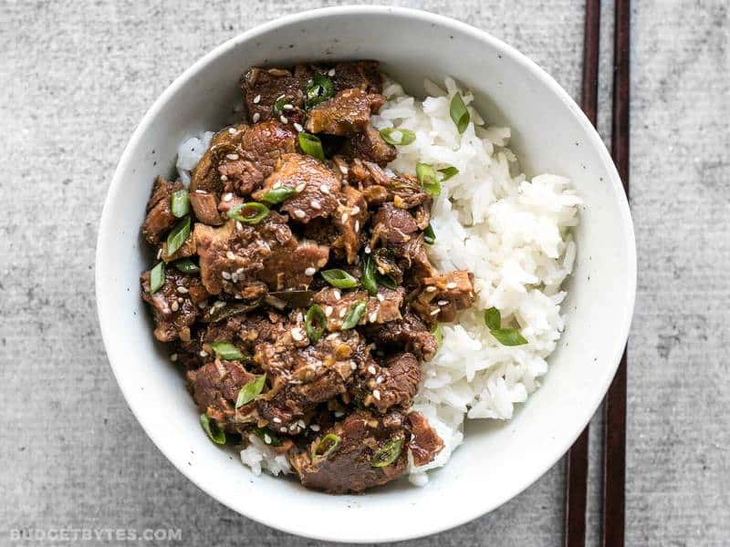 This tender, rich, and flavorful Slow Cooker Sesame Beef is extremely versatile and only requires a few ingredients that can be found at most grocery stores. BudgetBytes.com Slow Cooker Sesame Beef in a bowl with rice and green onion, chopsticks on the side