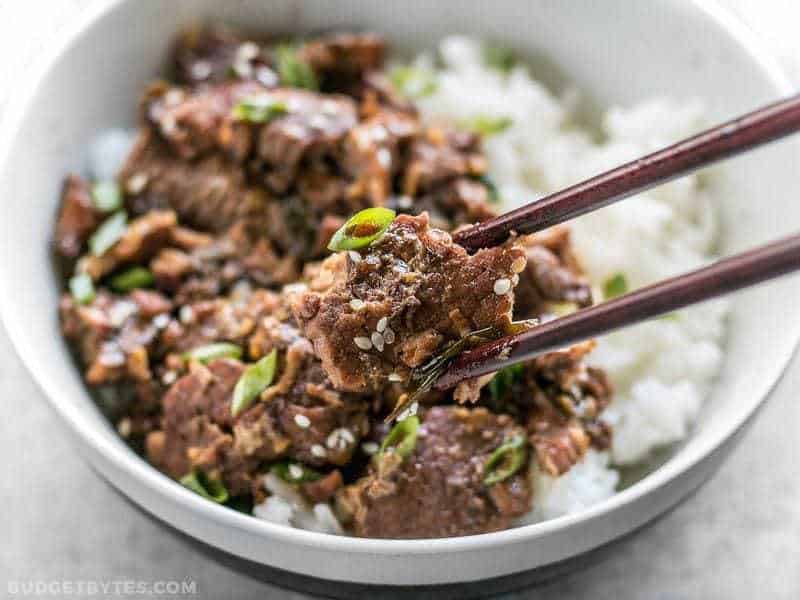 This tender, rich, and flavorful Slow Cooker Sesame Beef is extremely versatile and only requires a few ingredients that can be found at most grocery stores. BudgetBytes.com Close up of chopsticks picking up a piece of Slow Cooker Sesame Beef out of the bowl