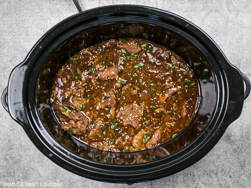 This tender, rich, and flavorful Slow Cooker Sesame Beef is extremely versatile and only requires a few ingredients that can be found at most grocery stores. BudgetBytes.com Slow Cooker Sesame Beef in the slow cooker garnished with green onion and sesame seeds
