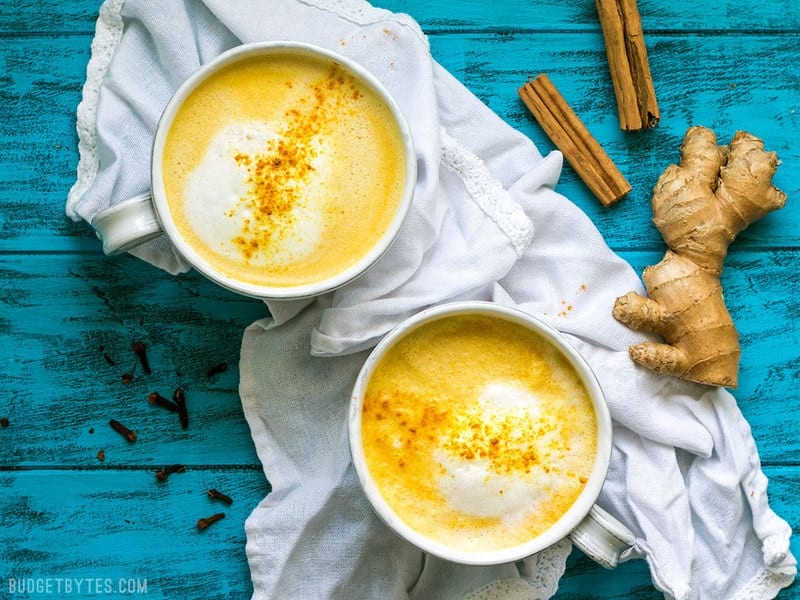 This Golden Chai is full of warm exotic spices, earthy turmeric, and just a hint of natural honey. It's warm, comforting, and perfect for fall! BudgetBytes.com Two mugs of Golden Chai with cinnamon sticks and a ginger root on the side