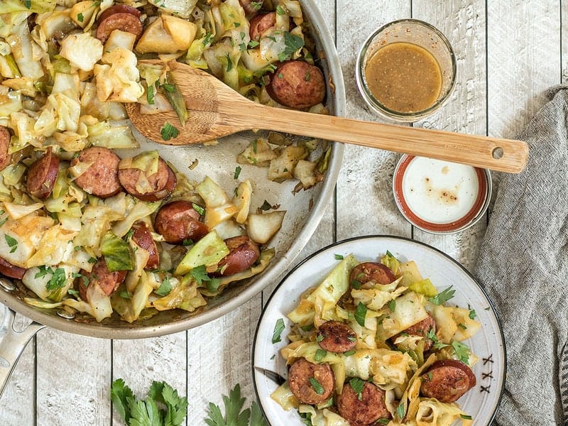 This Kielbasa and Cabbage Skillet is a fast and hearty weeknight dinner full of fiber and flavor! Serve with noodles or as is for a lower carb meal. Budgetbytes.com Kielbasa and Cabbage Skillet being served onto a plate from the skillet, with mustard vinaigrette in a jar on the side.