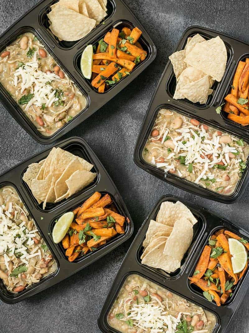 This White Chicken Chili Meal Prep includes a slow cooker chili with Cumin Lime Roasted Sweet Potatoes and tortilla chips for dipping! BudgetBytes.com Four White Chicken Chili Meal Prep containers scattered on a surface