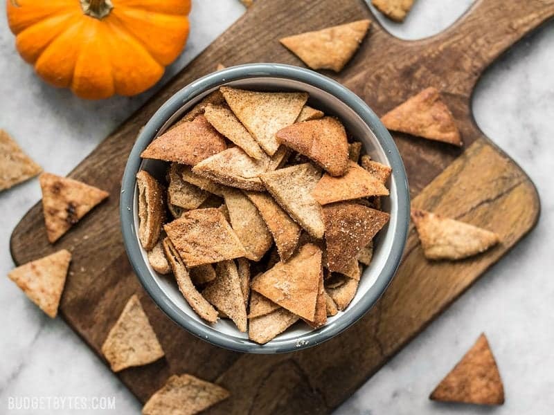 These Cinnamon Pita Crisps are a tasty little snack that's perfect for fall. Dip them into your favorite sweet treat or eat them on their own. BudgetBytes.com Finished Cinnamon Pita Crisps in a bowl next to a pumpkin
