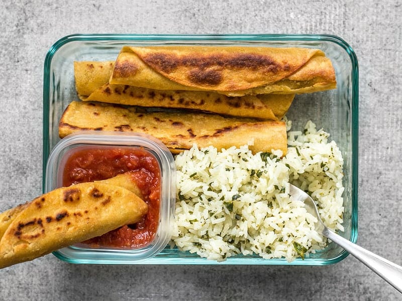 Creamy Black Bean Taquitos pair with tangy Cilantro Lime Rice for a simple and satisfying meal prep. BudgetBytes.com One Creamy Black Bean Taquito being dipped into salsa in the meal prep container