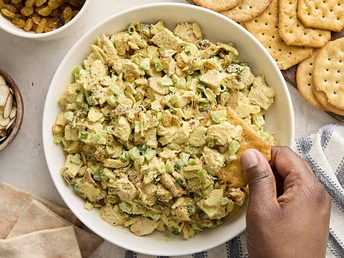 A hand dipping a cracking into a bowl of curry chicken salad.