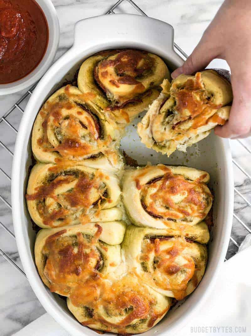 These Pesto Pizza Rolls are a fun way to change up your pizza routine with swirls of cheese, pesto, and pepperoni, plus lots of crispy edges! BudgetBytes.com A hand taking a Pesto Pizza Roll out of the baking dish