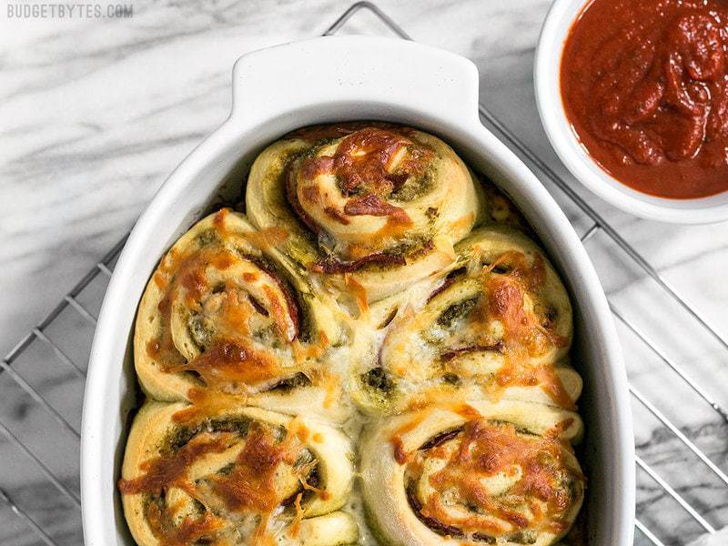 These Pesto Pizza Rolls are a fun way to change up your pizza routine with swirls of cheese, pesto, and pepperoni, plus lots of crispy edges! BudgetBytes.com Baked Pesto Pizza Rolls in the baking dish on a cooling rack with pizza sauce on the side
