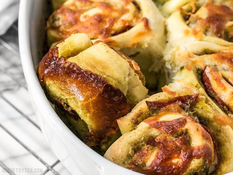 These Pesto Pizza Rolls are a fun way to change up your pizza routine with swirls of cheese, pesto, and pepperoni, plus lots of crispy edges! BudgetBytes.com Close up of a Pesto Pizza Roll turned sideways in the baking dish
