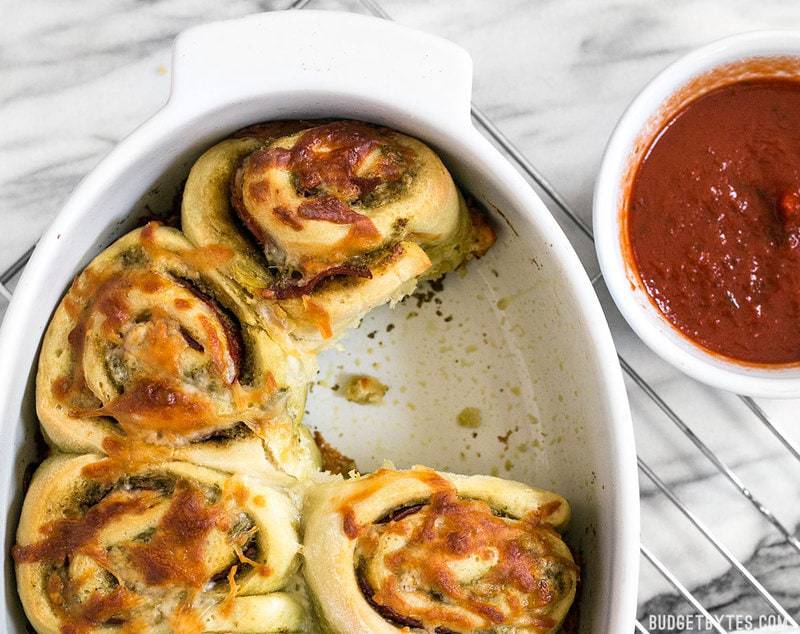 These Pesto Pizza Rolls are a fun way to change up your pizza routine with swirls of cheese, pesto, and pepperoni, plus lots of crispy edges! BudgetBytes.com Close up of the casserole dish of Pesto Pizza Rolls with one missing