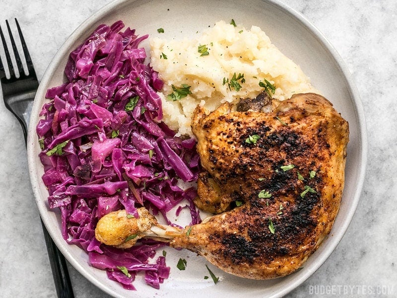 Braised Red Cabbage is an easy, cost efficient, and healthful side for your comforting winter meals. Step by step photos. Budgetbytes.com Braised Red Cabbage on a plate with a roasted chicken leg and mashed potatoes.