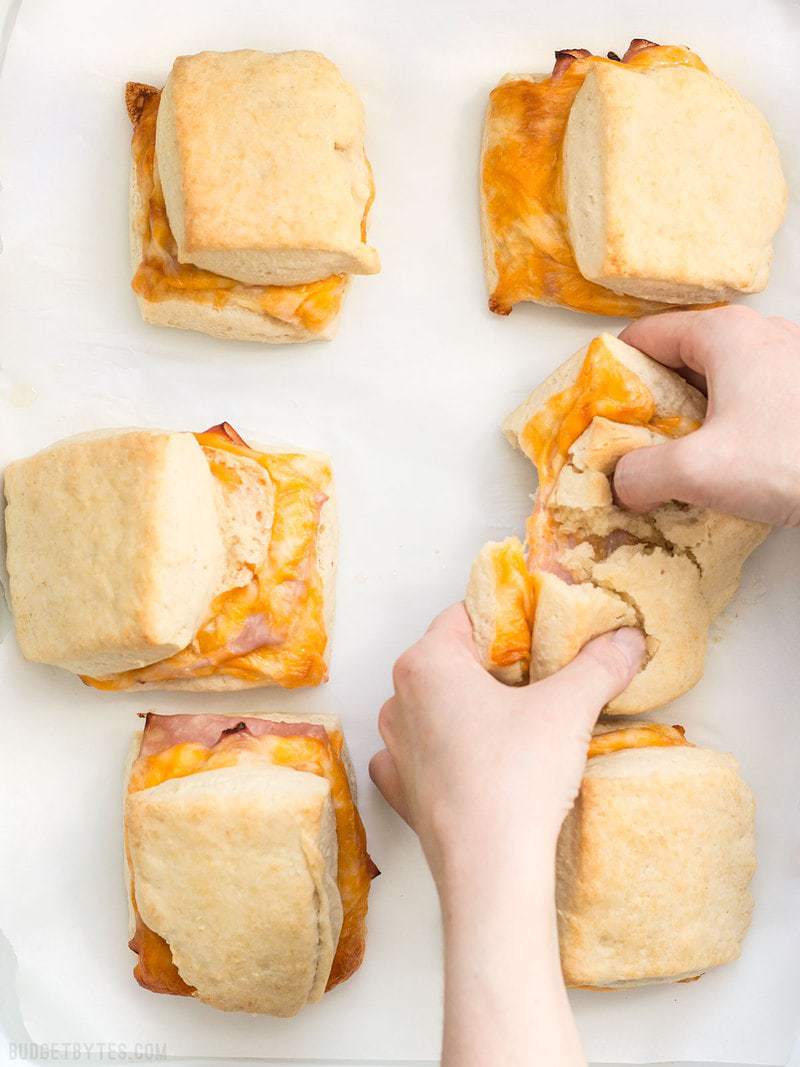 These Ham and Cheese Biscuits are a super indulgent, yet super easy treat for your weekend breakfast or to serve guests. BudgetBytes.com Six Ham and Cheese Biscuits on parchment, a hand tearing one in half.