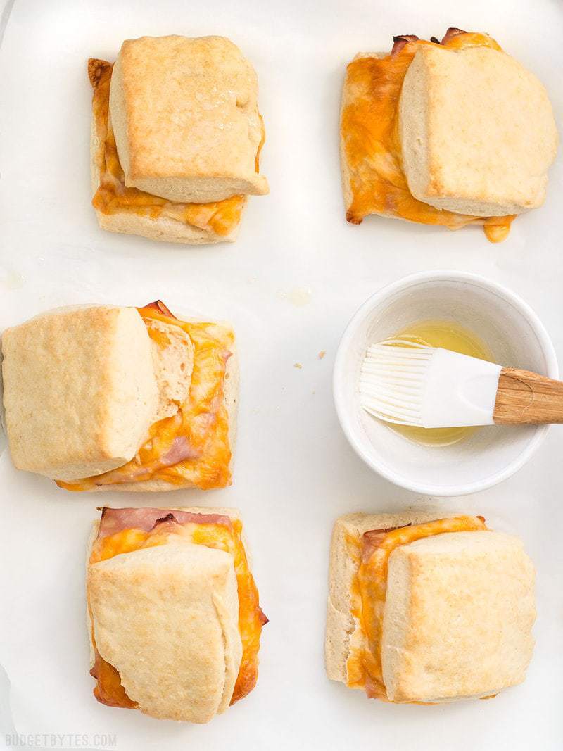These Ham and Cheese Biscuits are a super indulgent, yet super easy treat for your weekend breakfast or to serve guests. BudgetBytes.com Six Ham and Cheese Biscuits being brushed with melted butter