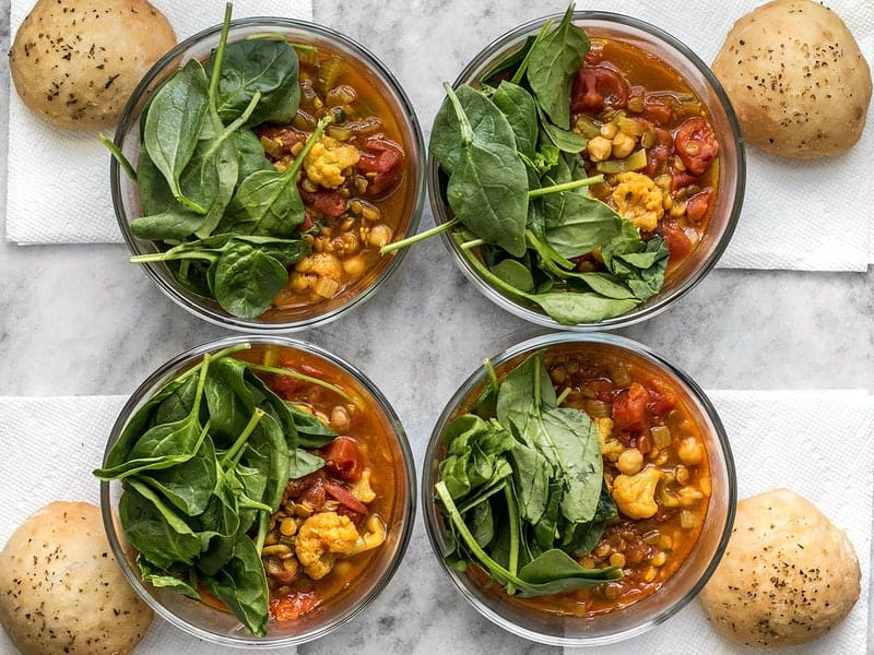 This Moroccan Lentil and Vegetable Stew meal prep is full of warm spices, homemade rolls, and an extra handful of spinach to make an absolutely killer meal. BudgetBytes.com Four Moroccan Lentil and Vegetable Stew meal prep bowls with spinach and focaccia rolls on the side