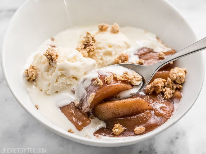 Slow Cooker Hot Buttered Apples are an easy and versatile dessert that is perfect for using up your glut of autumn apples! BudgetBytes.com A bowl of Slow Cooker Hot Buttered Apples topped with ice cream and granola, being eaten with a spoon.