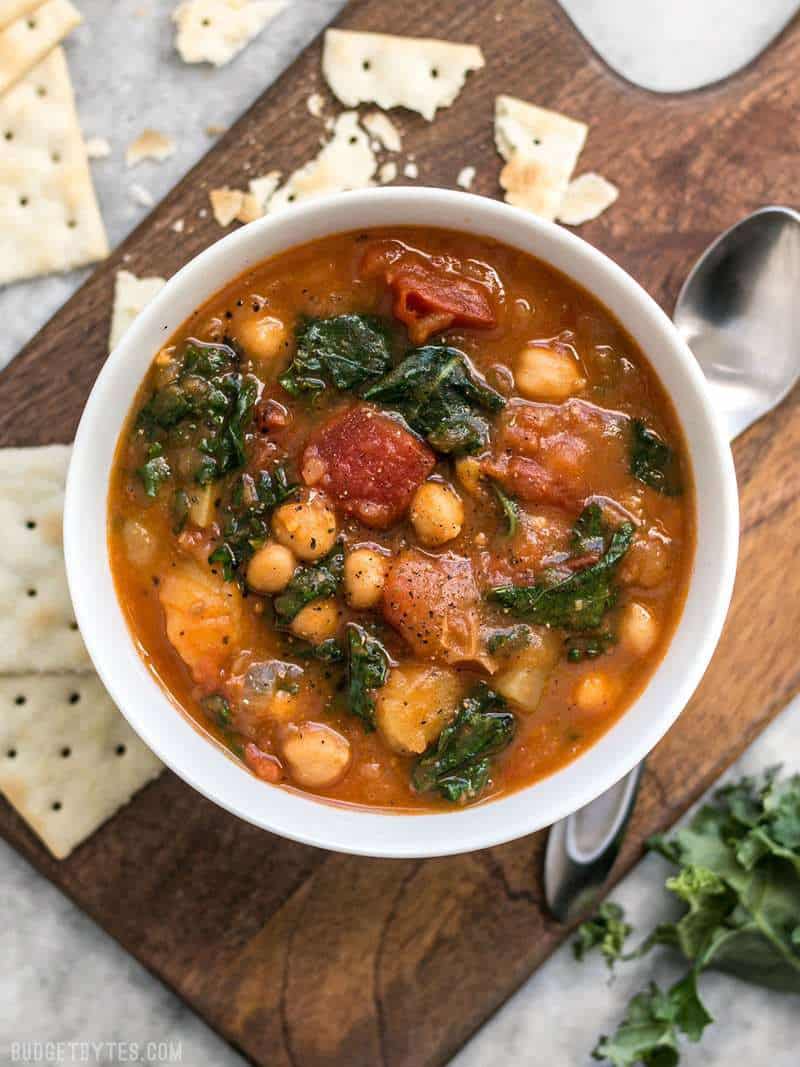 This Smoky Potato Chickpea Stew is a hearty and filling plant-based dish that will keep you full and warm this winter! BudgetBytes.com A bowl of Smoky Potato Chickpea Stew on a wooden cutting board surrounded by saltine crackers.