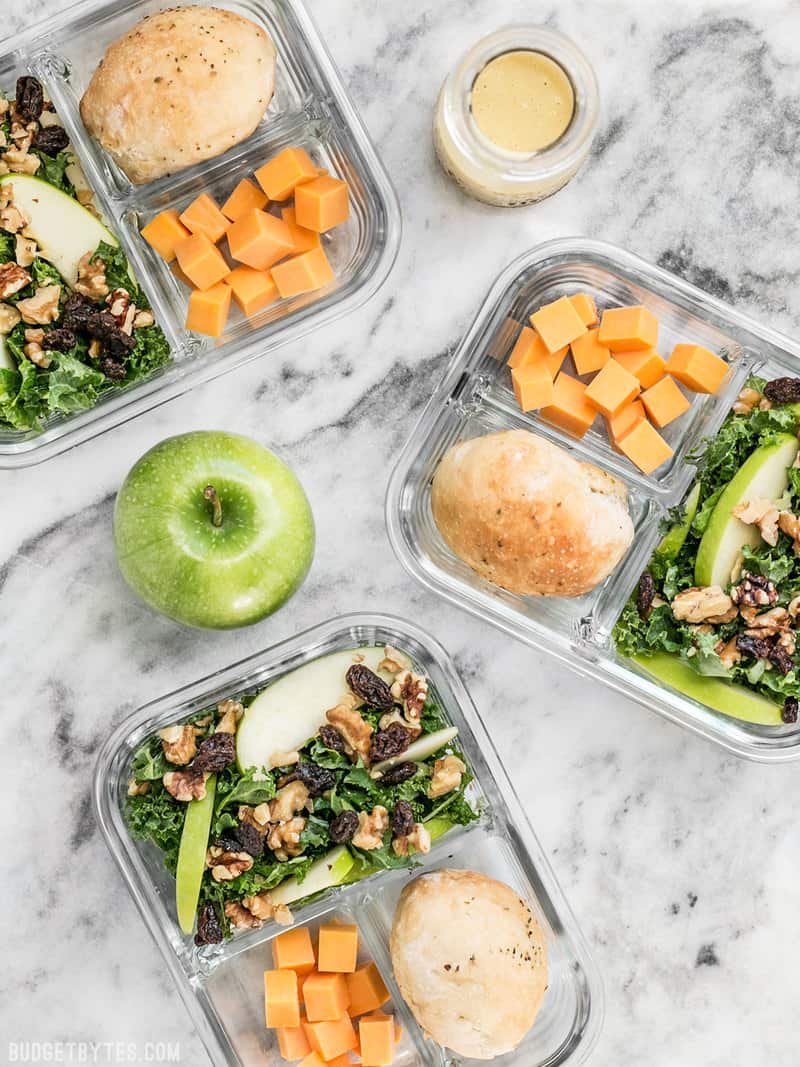 This Apple Dijon Kale Salad Meal Prep is full of texture and bright flavors, and is sure to keep you full for hours. BudgetBytes.com This Apple Dijon Kale Salad Meal Prep containers scattered, with an apple and dressing near by.