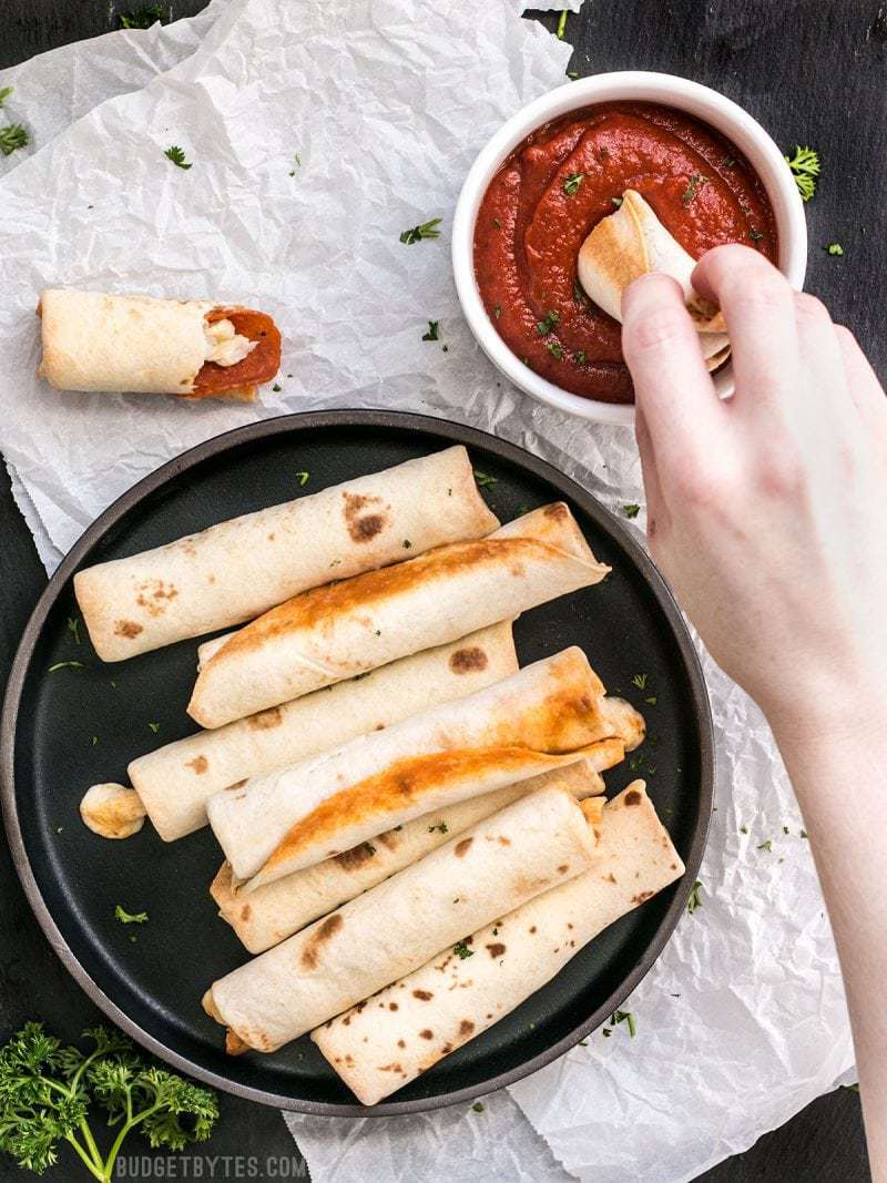 Forget frozen pizza rolls! These Pizza Roll Ups are the perfect last minute appetizer or indulgent snack on busy days! BudgetBytes.com A plate full of Pizza Roll Ups and a hand dipping one into a bowl of pizza sauce