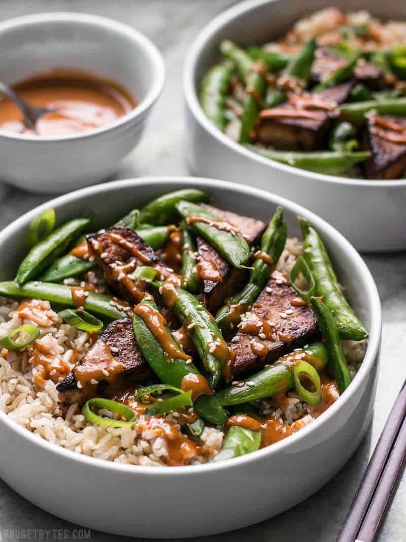 These vegan Soy Marinated Tofu Bowls are full of rich flavors and plenty of texture to keep your taste buds happy and your belly full. BudgetBytes.com Two Soy Marinated Tofu Bowls with spicy peanut sauce in the background