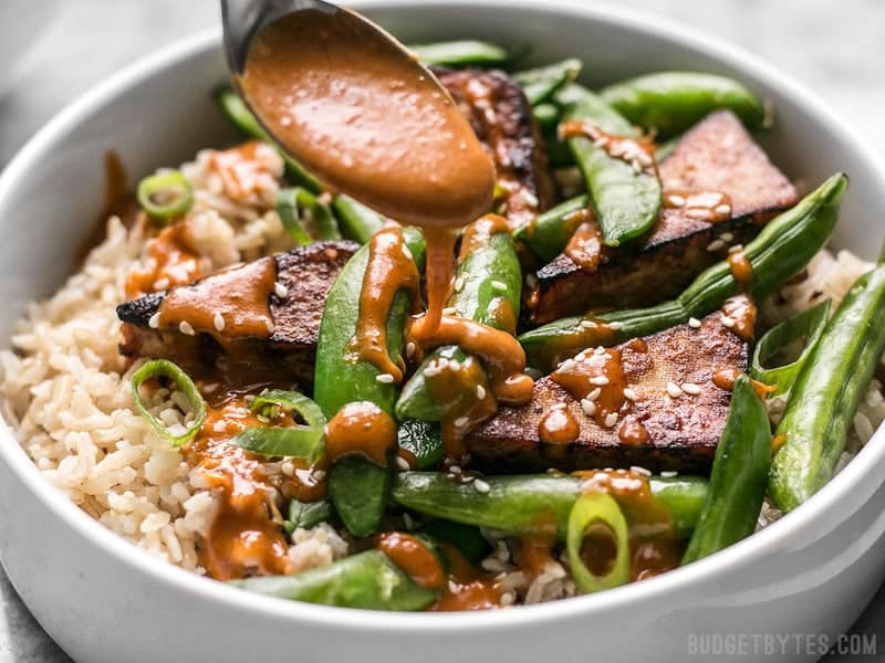 These vegan Soy Marinated Tofu Bowls are full of rich flavors and plenty of texture to keep your taste buds happy and your belly full. BudgetBytes.com Spicy Peanut Sauce being drizzled on a Soy Marinated Tofu Bowl